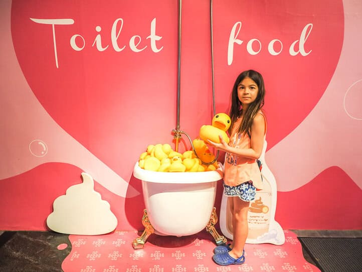 Una niña parada junto a una pared pintada de rosa oscuro y claro, con media bañera llena de patitos de goma amarillos que sobresalen de la pared y las palabras "Comida de baño" en la pared.