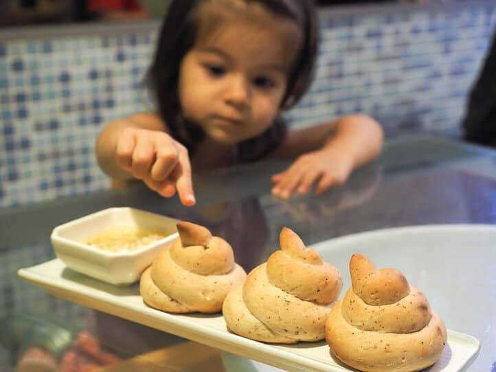 Una niña extiende la mano para pinchar uno de los tres bollos con forma de caca que hay en una bandeja blanca larga y delgada sobre la mesa frente a ella.