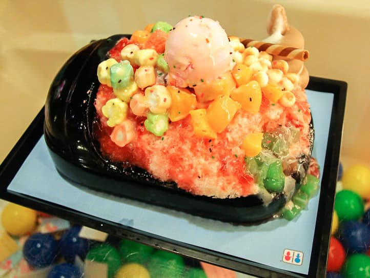 Un postre abundante de hielo raspado servido en un recipiente negro en forma de inodoro, con varios dulces de colores, malvaviscos, galletas y helado encima, todo bañado en salsa de fresa.