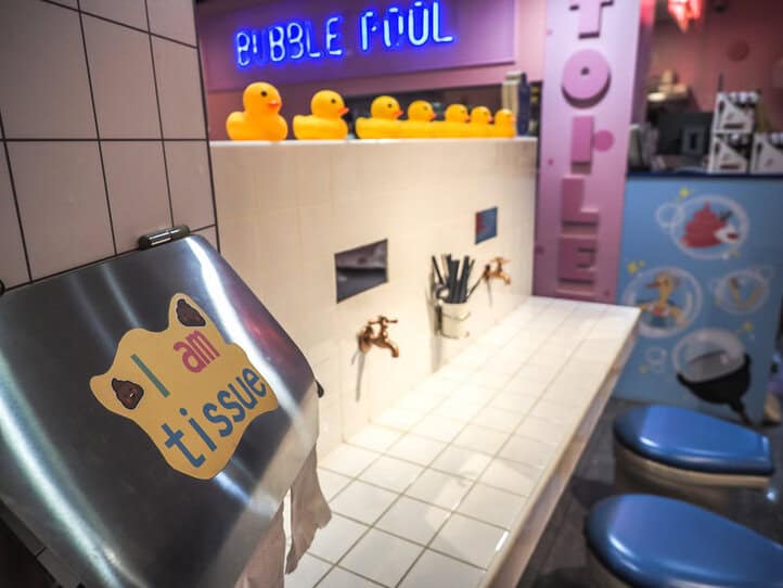 Dentro de un restaurante, con dos sillas en un mostrador embaldosado como un baño, con dos grifos en la pared, un dispensador de pañuelos que dice "Soy un pañuelo" y una barra detrás con las palabras de neón "Bubble Pool" encima.