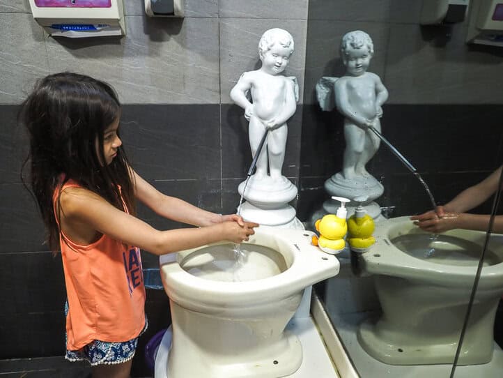 Una niña se lava las manos en un baño, con el agua saliendo de una estatua de un niño, como si estuviera orinando.