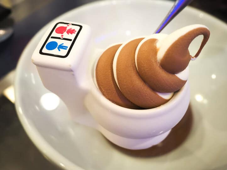 Un plato blanco con forma de inodoro que contiene helado suave de chocolate y vainilla, con símbolos de un niño azul y una niña rosa sobre él.