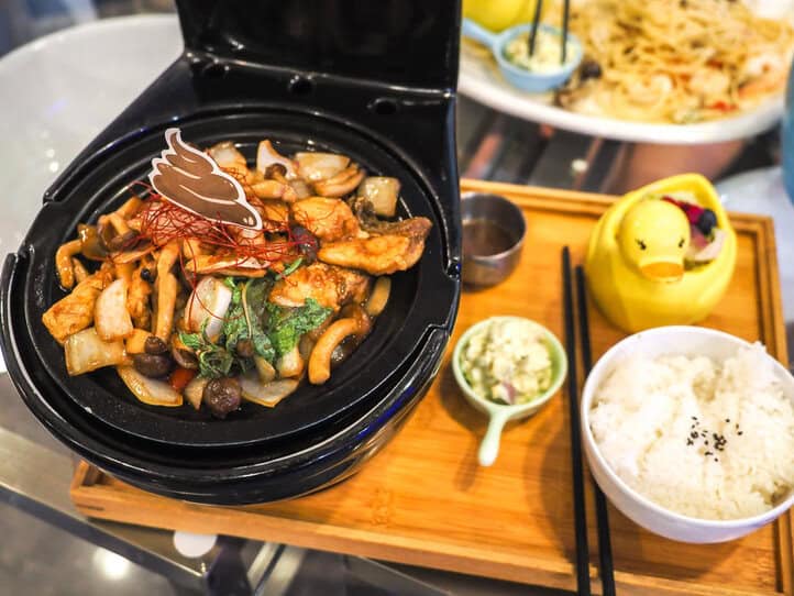Un inodoro en miniatura negro lleno de pescado frito y champiñones, sobre una bandeja de madera con algunas guarniciones que incluyen un tazón de arroz y un patito de goma relleno de ensalada.