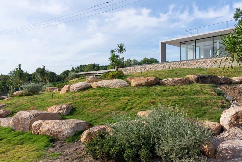 Modern hillside house with glass walls and stone accents, surrounded by lush greenery and large rocks, showcasing contemporary architecture and landscape design.