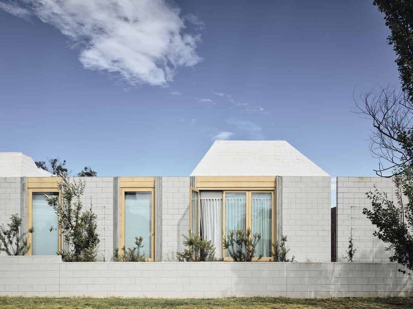 Modern minimalist residential architecture with clean lines, white brick facade, large glass windows, and wooden accents, set against a vibrant blue sky and surrounded by greenery.