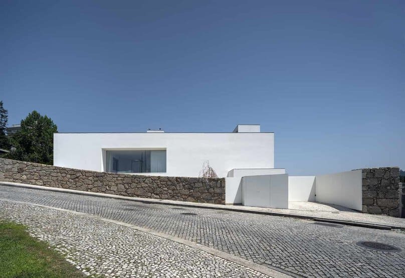 Modern minimalist white house with clean lines and large glass windows, situated on a sloped cobblestone street with stone walls, showcasing contemporary architecture and innovative design elements.