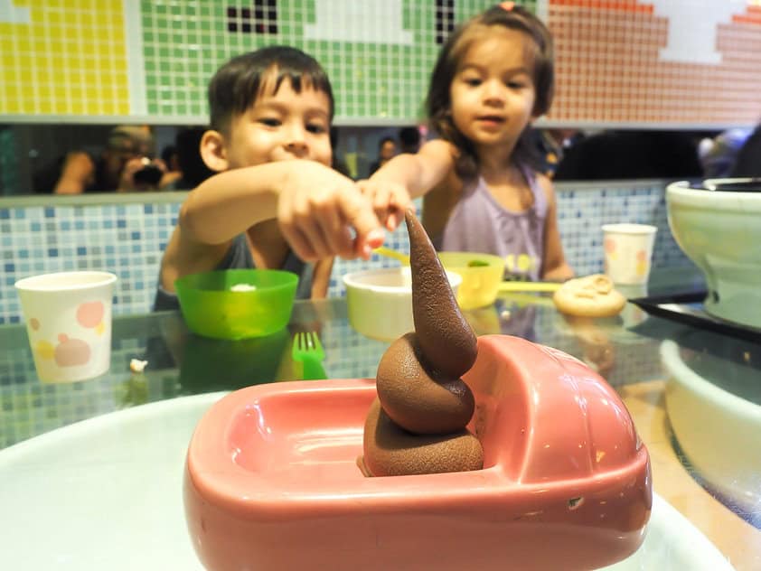 Dos niños en un restaurante con temática de baño, ambos señalando una pila de helado de chocolate suave que parece caca y servido en un plato rosa que parece un inodoro.