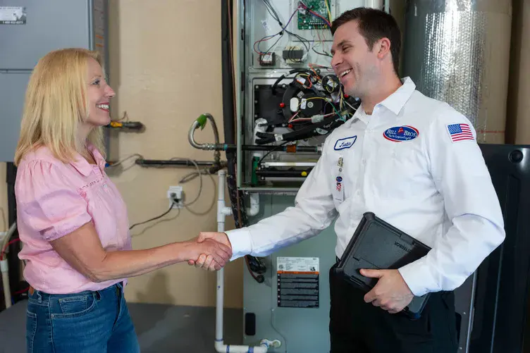 A woman greets a friendly, uniformed Bell Brothers technician—background-checked and ready with upfront pricing—in front of a home heater.