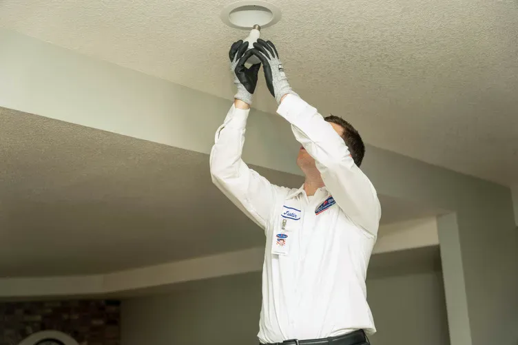 A Bell Brothers technician, uniformed and gloved, safely installs a ceiling light, assuring upfront pricing and No Surprise Guarantee.