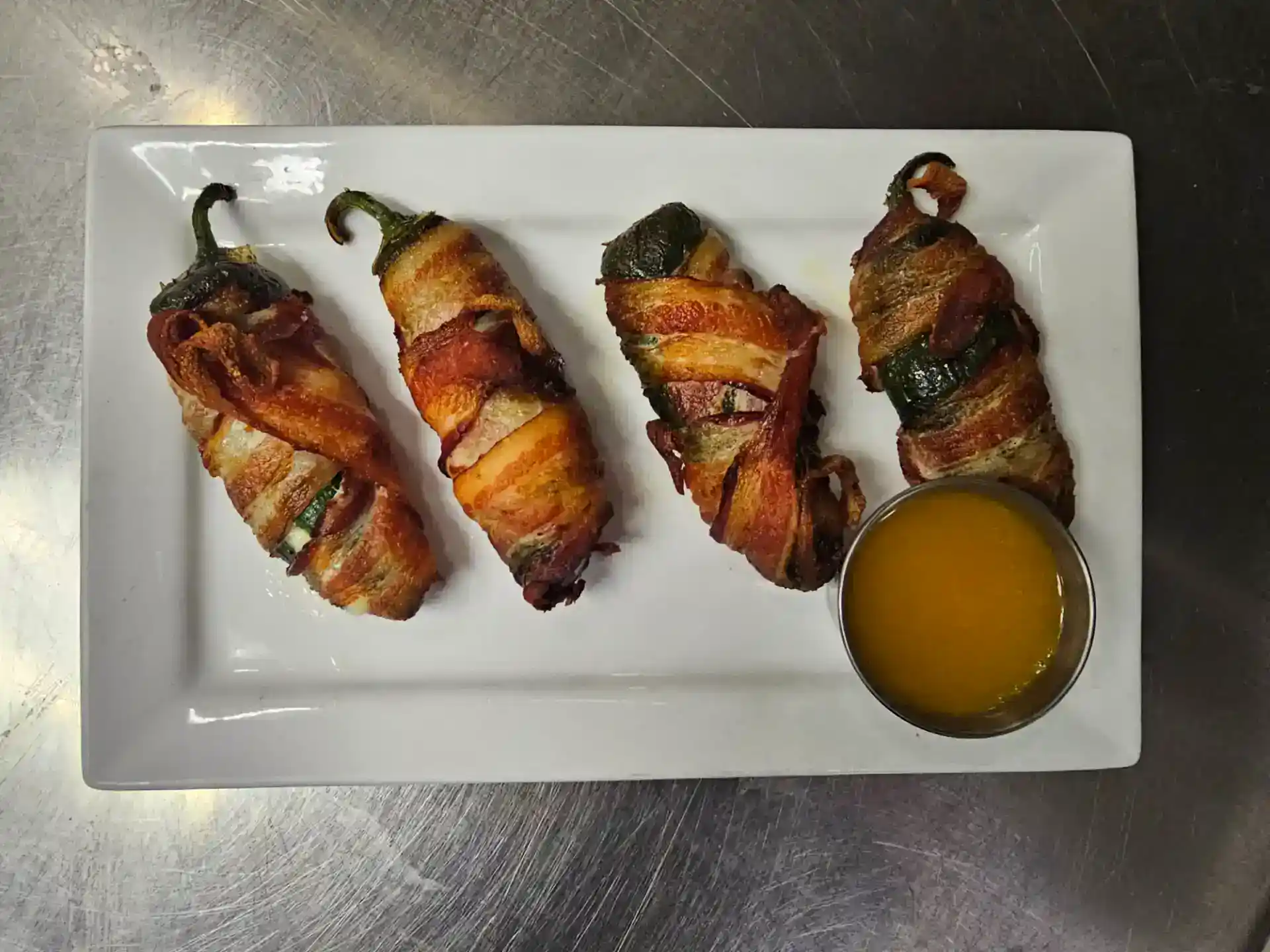 Bacon-wrapped jalapeño poppers with cheese on a white plate with dipping sauce.