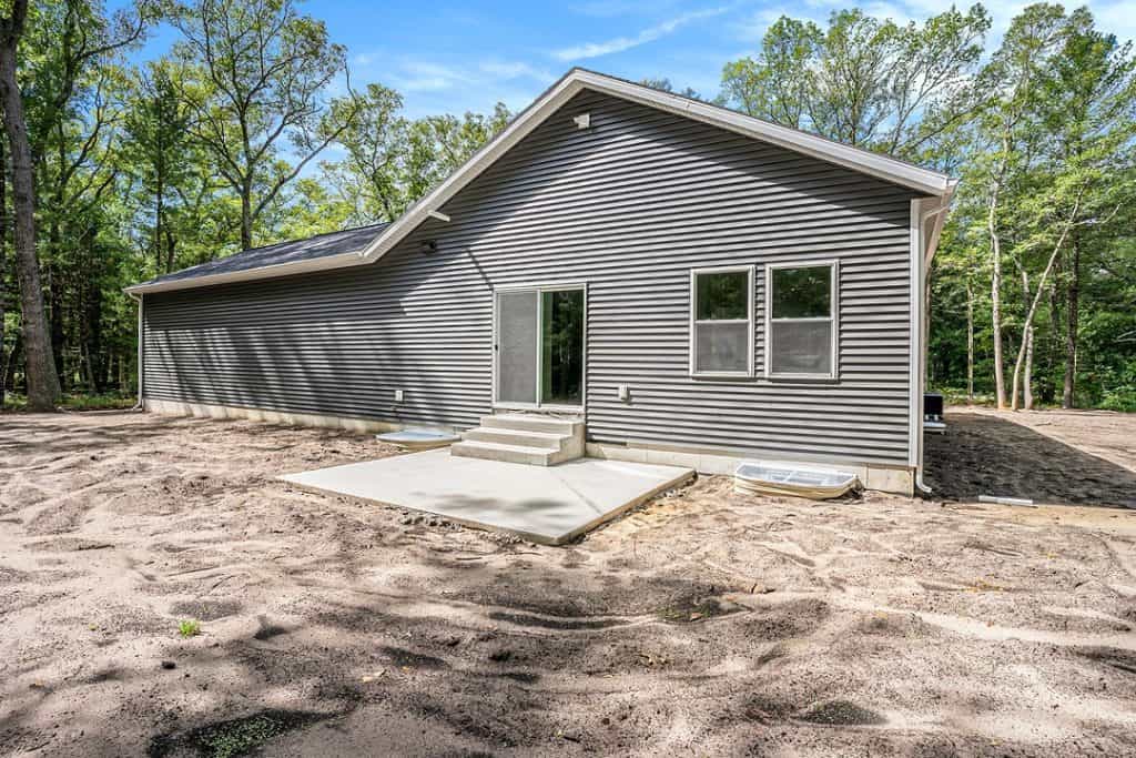 Newly built custom home with gray siding and large windows in a wooded area.