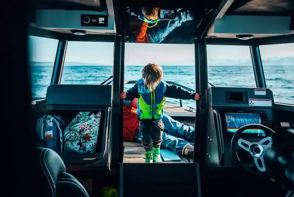 Child in life jacket steering boat on open water.