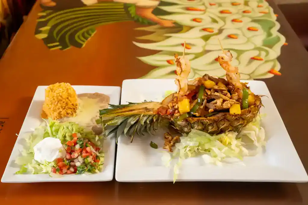 Seafood stuffed pineapple with vegetables and garnishes on a white plate.