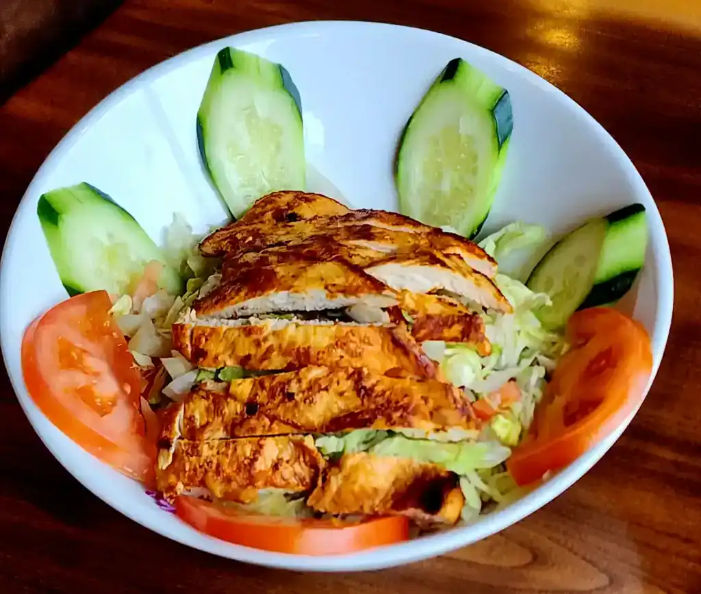 Grilled chicken salad with fresh cucumbers, tomatoes, and lettuce in a white bowl.