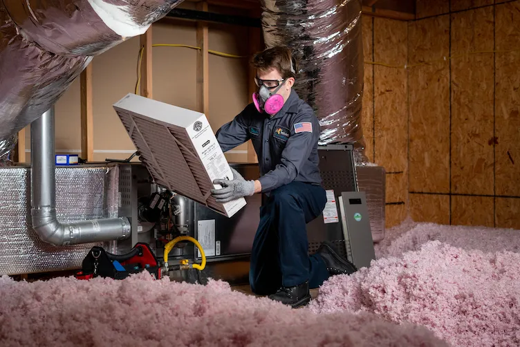 A friendly Fetch‑A‑Tech technician, equipped with a protective mask and gloves, carefully installs a new air filter in an HVAC system in a cozy Las Vegas attic lined with pink insulation—making sure your home stays comfortable and healthy for your family.