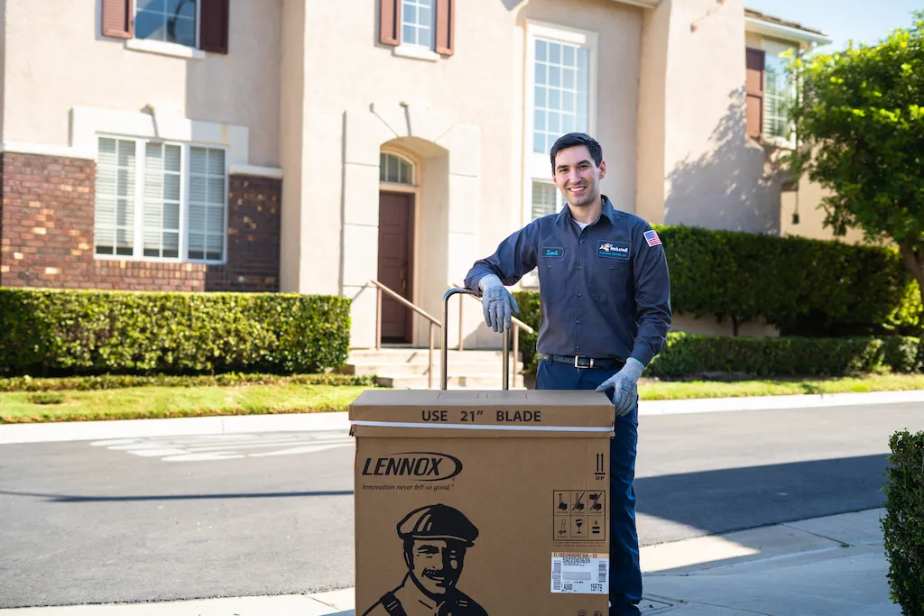 A friendly Fetch‑A‑Tech technician, dressed in uniform and wearing gloves, stands outside a Las Vegas home with a warm smile. He rests his hand on a large Lennox box on the sidewalk, ready to provide reliable and helpful service to ensure your family’s comfort.