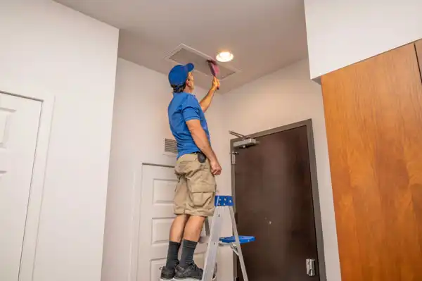 A friendly technician from Fetch‑A‑Tech stands on a step ladder, carefully cleaning a ceiling air vent with a brush to help keep a Las Vegas home comfortable and welcoming.