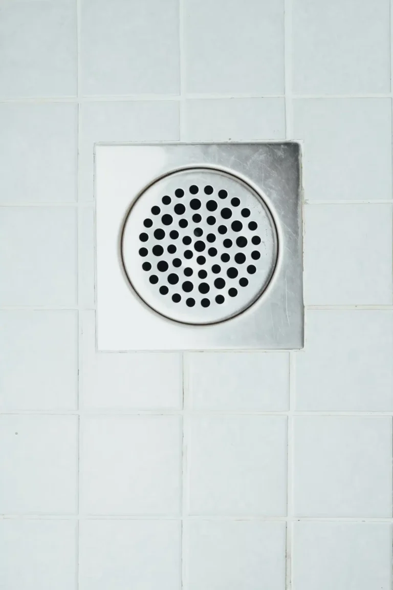 A metal floor drain cover with round holes is securely installed in clean, white tile flooring—showcasing the careful craftsmanship and reliable plumbing service Fetch‑A‑Tech provides to Las Vegas families.