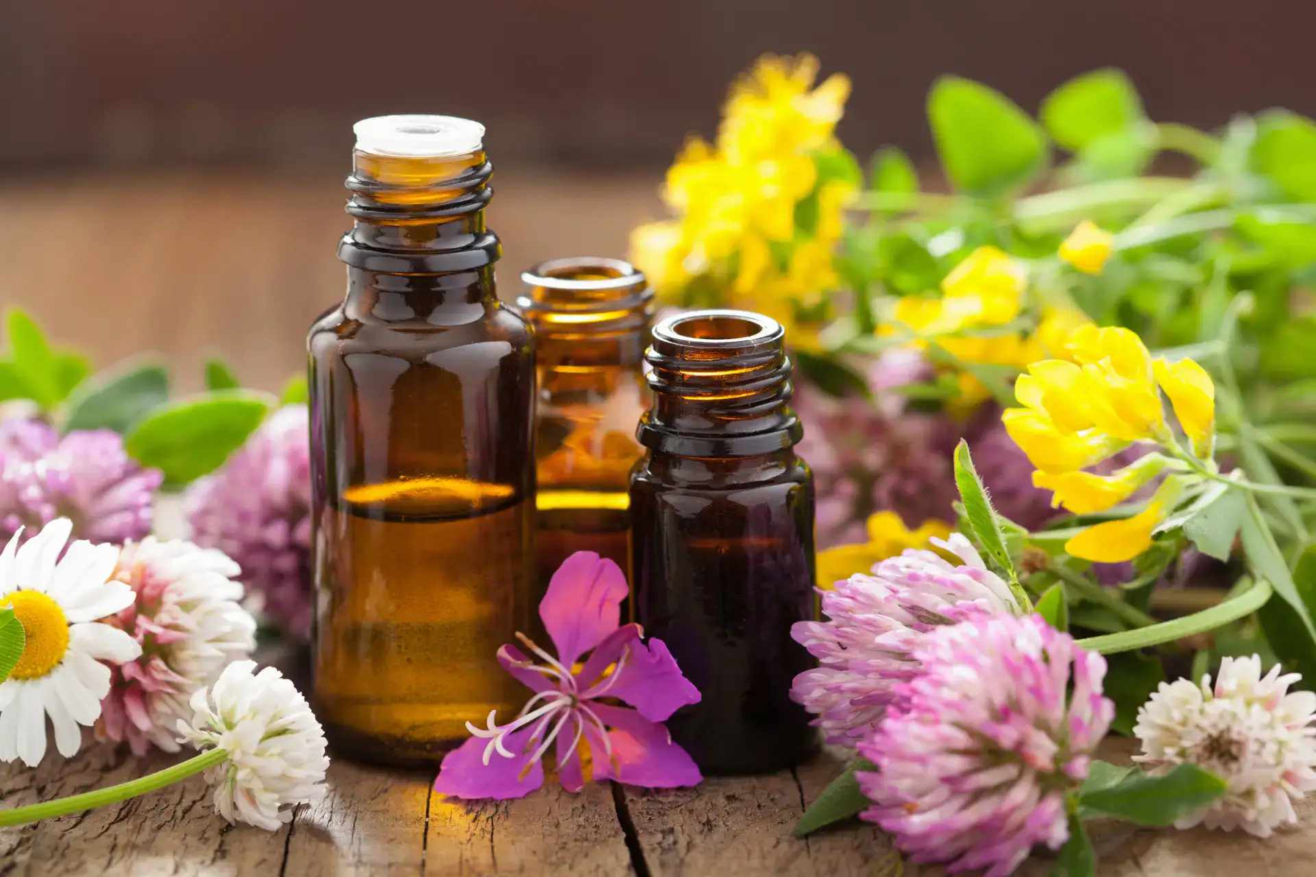 Three amber essential oil bottles surrounded by colorful flowers on a wooden surface.