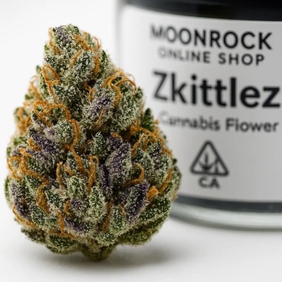 Close-up macro of a Zkittlez cannabis bud covered in frosty trichomes with purple hues and orange pistils, jar label softly blurred in the background — Moonrock Online Shop.