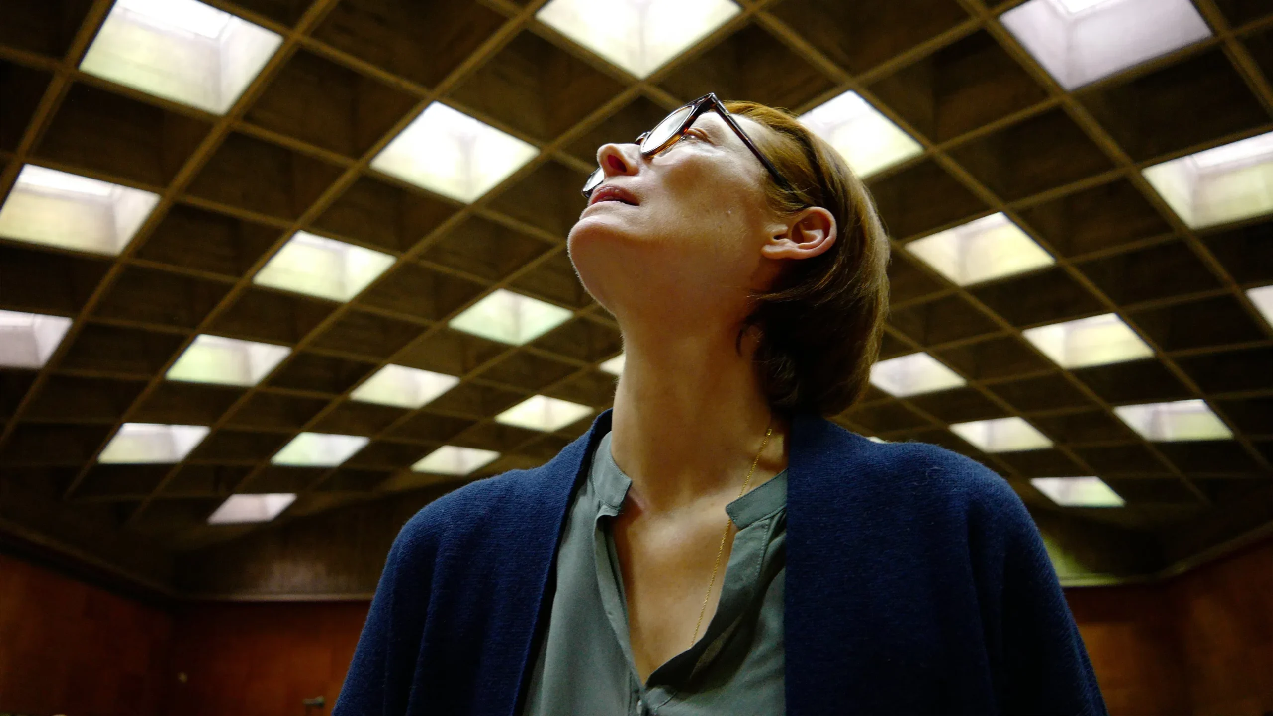 A person with short hair and glasses looks upward, standing indoors under a large grid-patterned ceiling with square skylights. The lighting creates a geometric pattern overhead.
