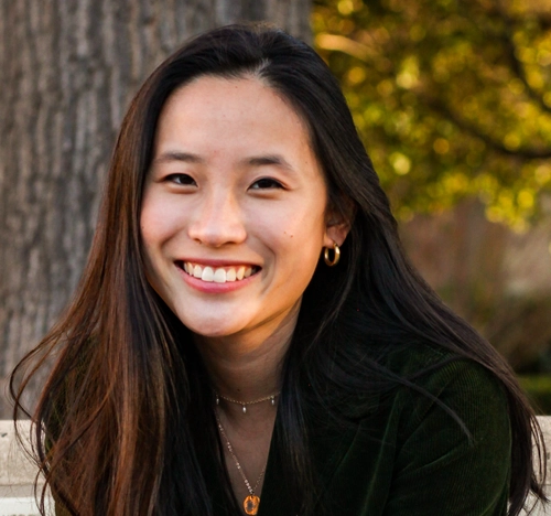 Christina Li, a woman with long dark hair, smiles while sitting outdoors. She wears gold hoop earrings, layered necklaces, and a dark top. Trees and greenery are visible in the blurred background.