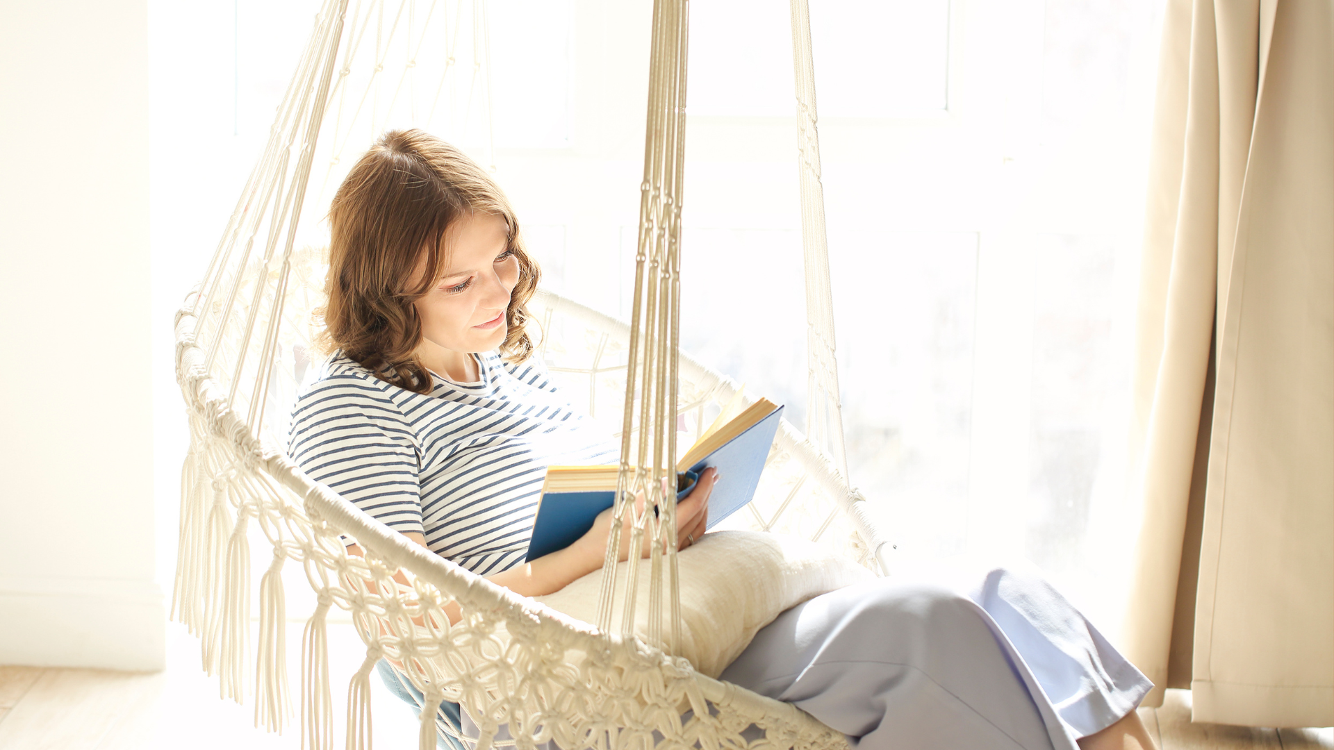 A woman sitting in a hammock smiling as she reads a book, clearly practicing self-care while working from home