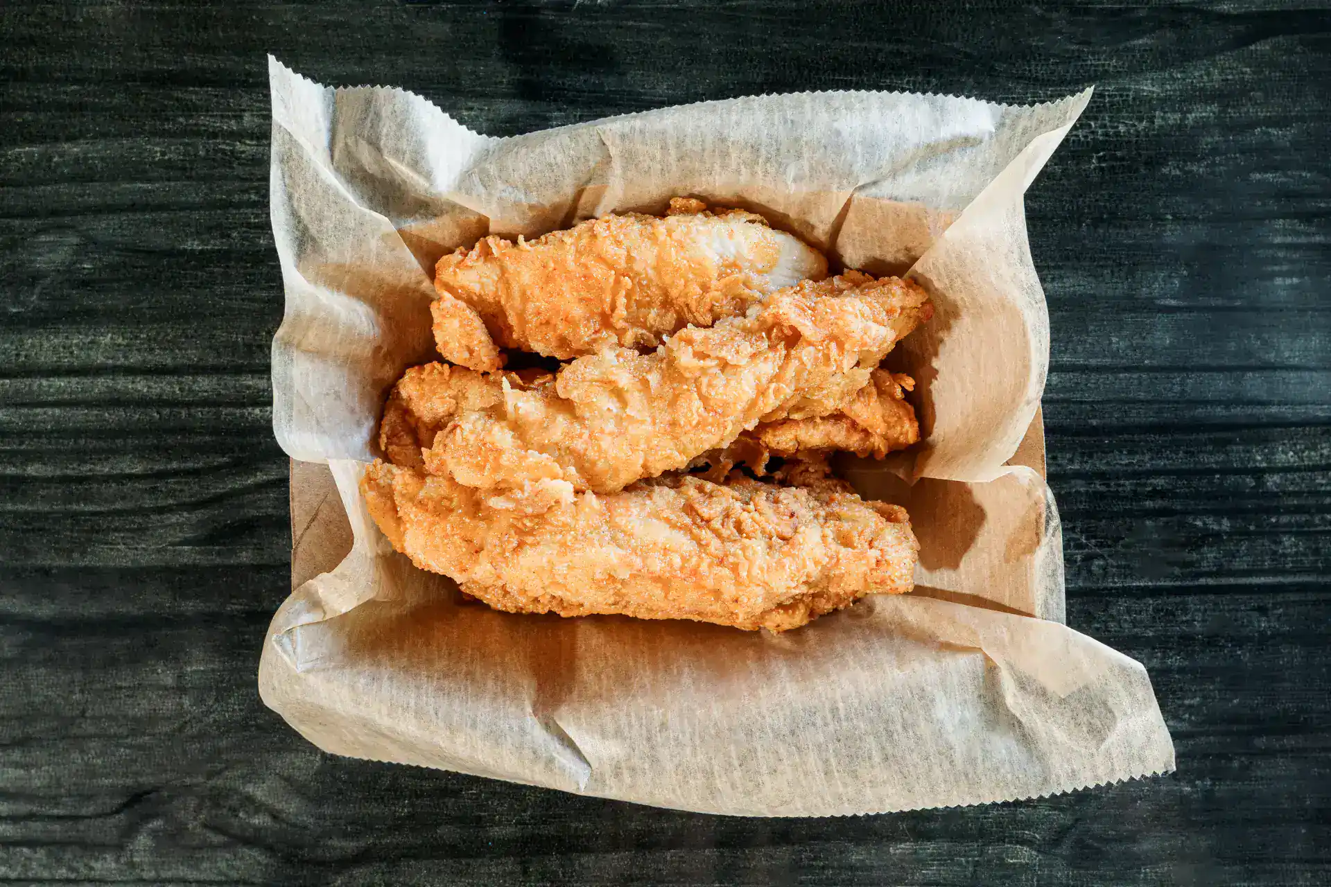 Golden fried chicken strips on parchment paper on dark wood table.