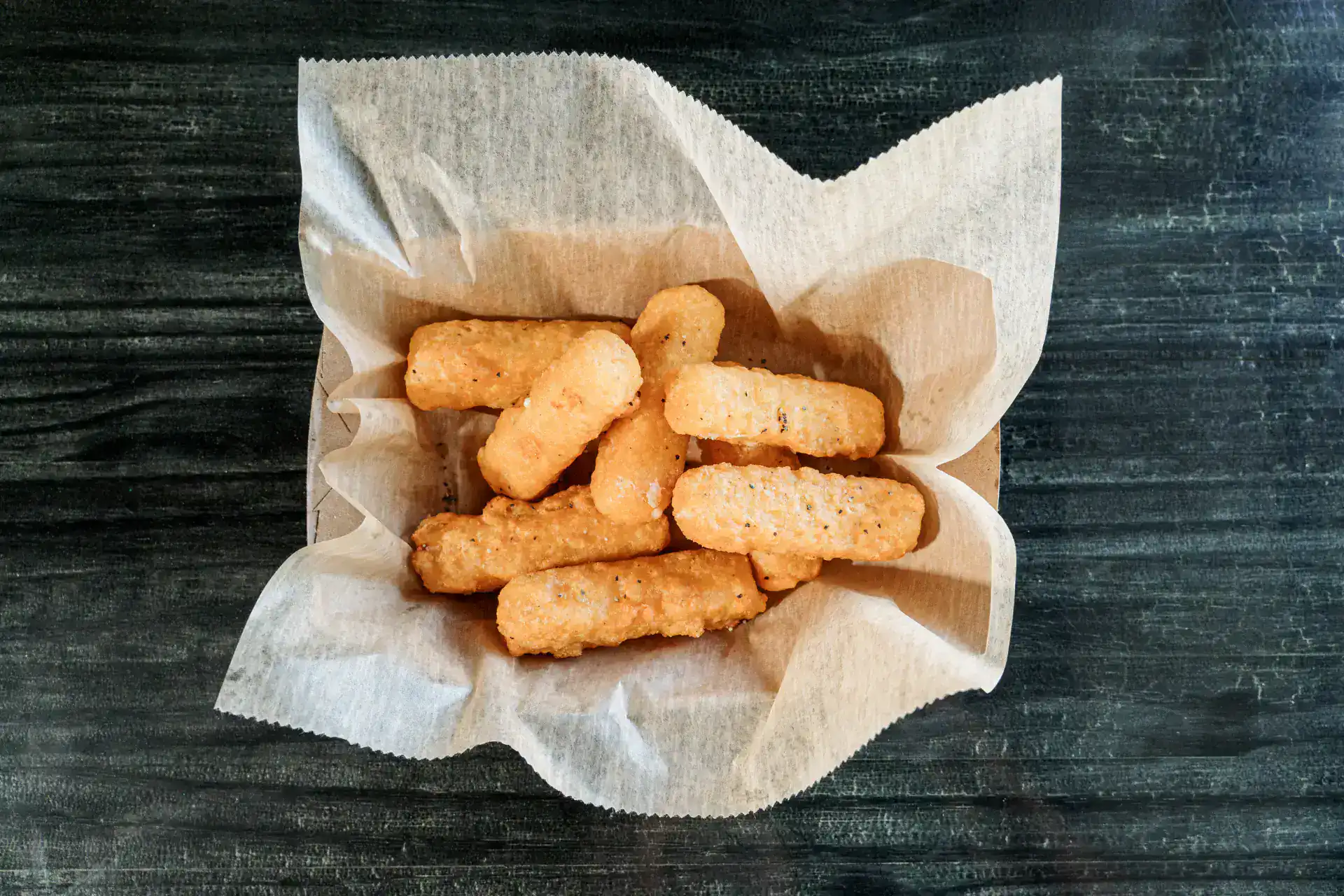 Golden fried cheese sticks on parchment paper on dark wooden surface.