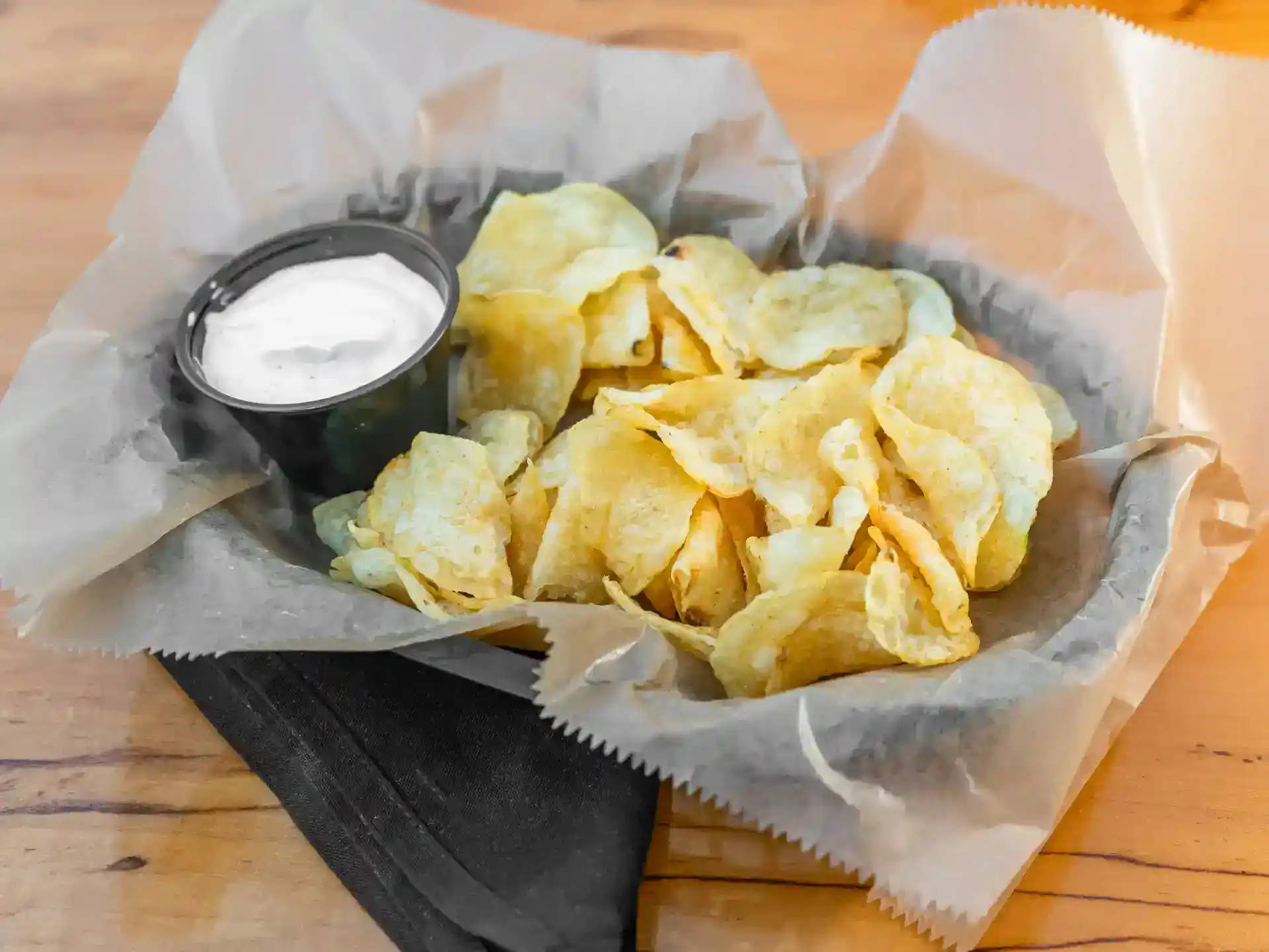 Potato chips with a side of sour cream in a black cup on parchment paper.