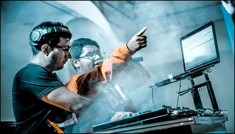 Two men work at a music production setup with a laptop; one wears headphones and uses equipment while the other points at the screen amid blue-tinted stage lighting and smoke.