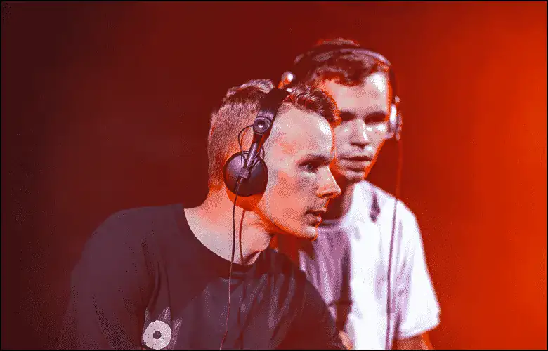 Two men wearing headphones stand side by side, focused on DJ equipment, illuminated by red stage lighting.