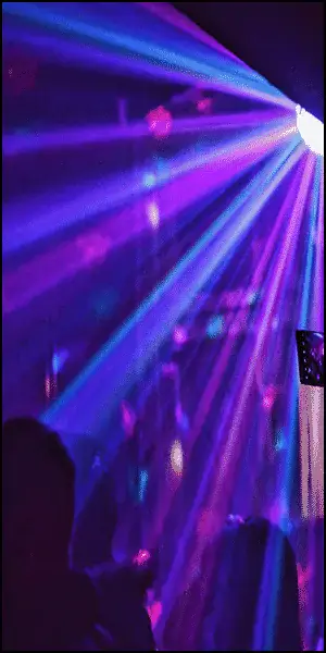 Purple and blue laser lights shine through a dark room, illuminating silhouettes of people at what appears to be a party or concert.