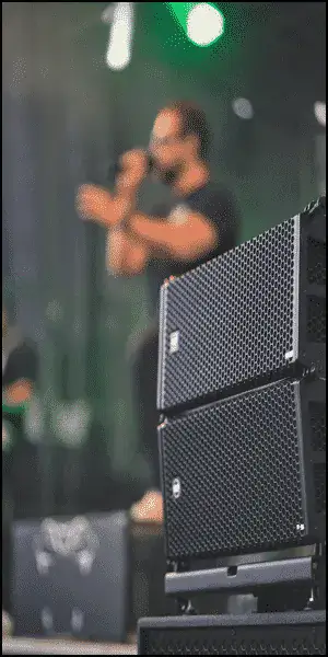 A stack of black speakers on an outdoor stage with a blurred person holding a microphone in the background.