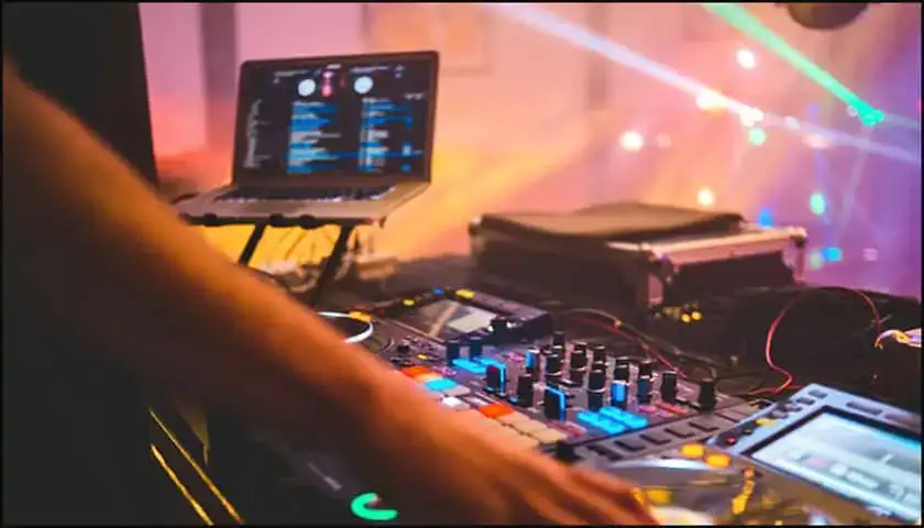 A person operates a DJ mixer and turntables with a laptop displaying music software; colorful lights illuminate the background.