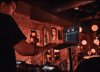 A DJ stands at a booth holding a laptop, with warm lighting and framed photos on the wall behind him in a dimly lit venue.