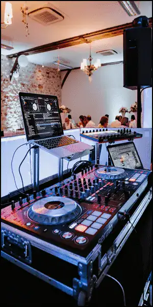 A DJ controller and laptop setup in a decorated event space with people and chandeliers visible in the background.