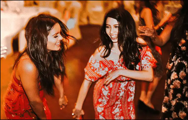 Two women in red dresses are smiling and dancing together at an indoor event with other people in the background.