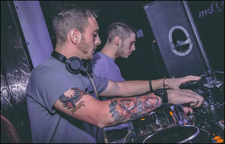 Two DJs stand side by side operating a mixer and turntables; one has tattoos on his arm and headphones around his neck.