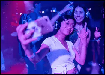 A group of young people wearing headphones pose for a selfie in a dimly lit, purple-toned party setting.