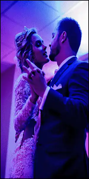 Bride and groom in formal attire share a close dance under colorful lighting, looking into each other's eyes.