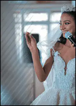 A bride in a white wedding dress holds a microphone and smiles, raising her hand while standing indoors.
