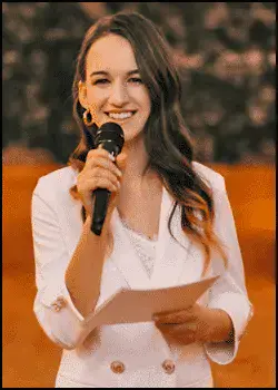 Woman in a white blazer holds a microphone and papers, smiling while speaking, with a blurred orange and dark background.