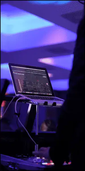 A DJ setup with a laptop displaying music software on a stand, illuminated by blue and purple lights in a dim indoor setting.
