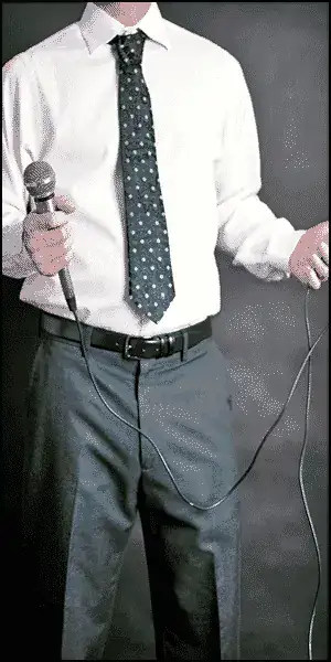 Man in a white shirt and tie holding a microphone with a long cord, standing against a plain dark background.