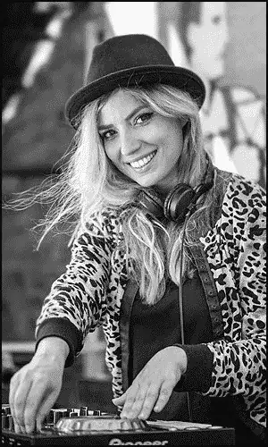A woman wearing a hat and leopard-print jacket stands at a DJ mixer, smiling at the camera with headphones around her neck.