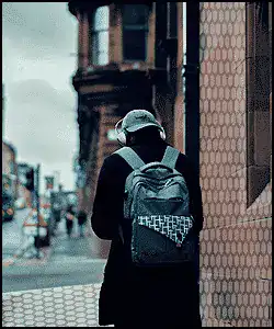 A person wearing headphones and a backpack walks along a city street near a brick building on a cloudy day.
