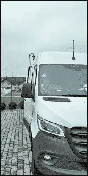 A white van is parked on a paved driveway, with a person visible in the driver's seat. The background shows houses and a cloudy sky.