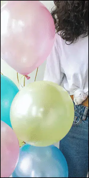 A person wearing a white shirt and blue jeans stands beside pastel-colored balloons, including pink, blue, and yellow.