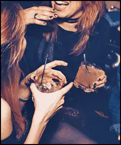 Two people sitting close together, both holding drinks, smiling and laughing in a dimly lit setting.